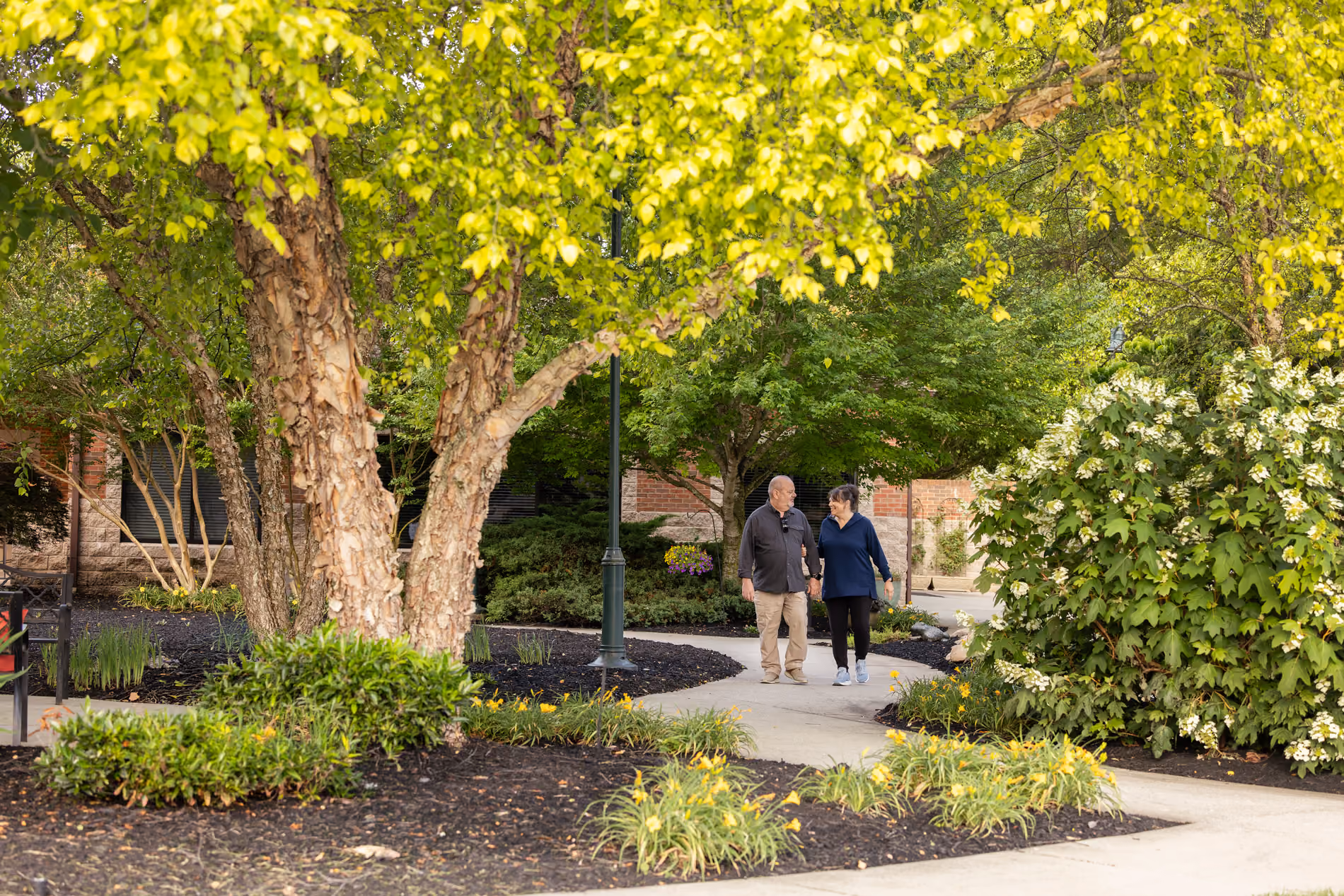 An older couple walking hand in hand on a garden path surrounded by green trees and flowers.