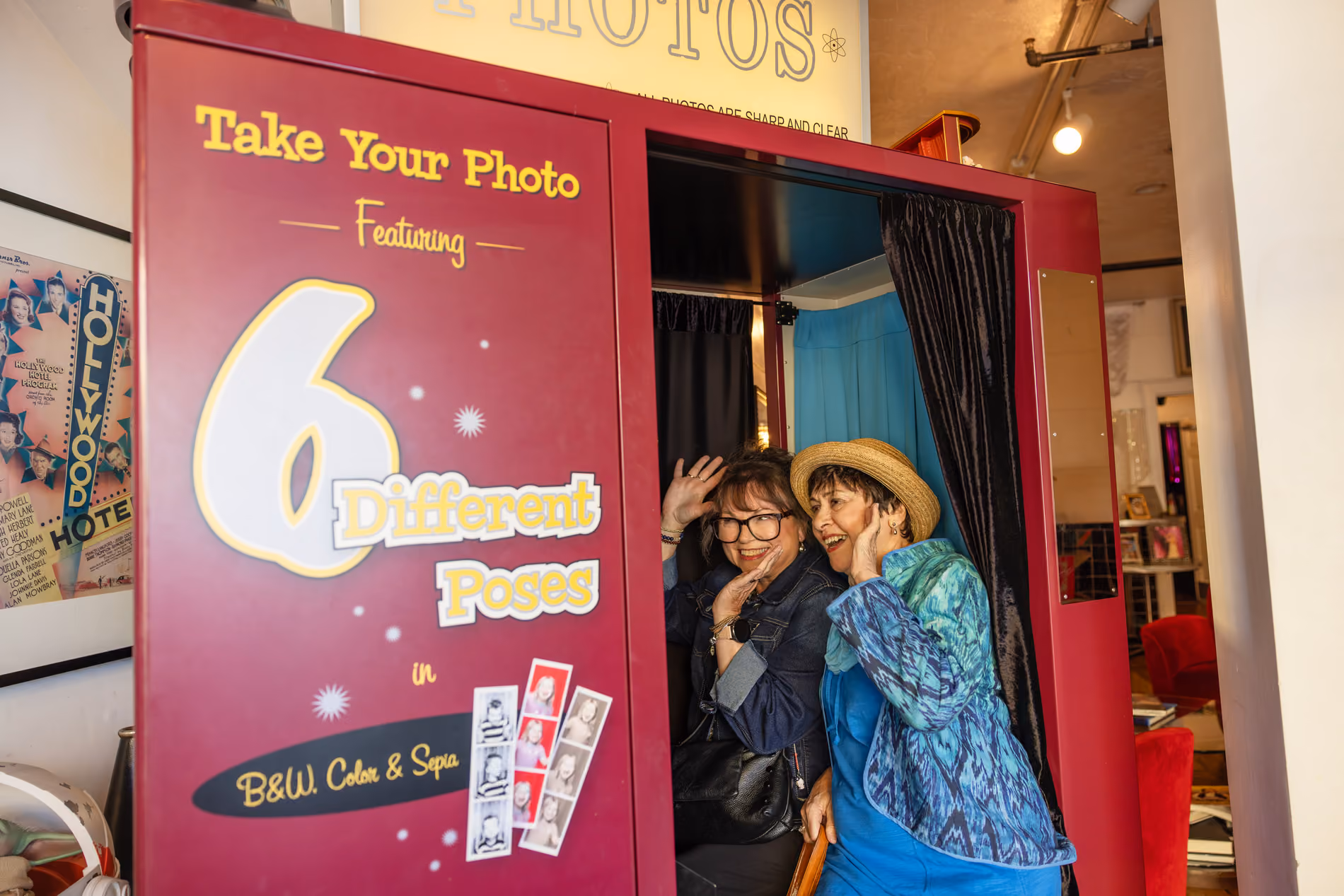 Two women posing and smiling inside a photo booth with red curtains and a denim jacket.