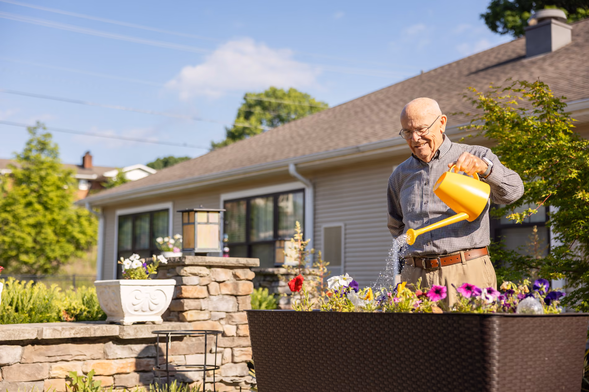 Elderly man watering colorful flowers in a garden planter outside a house on a sunny day.