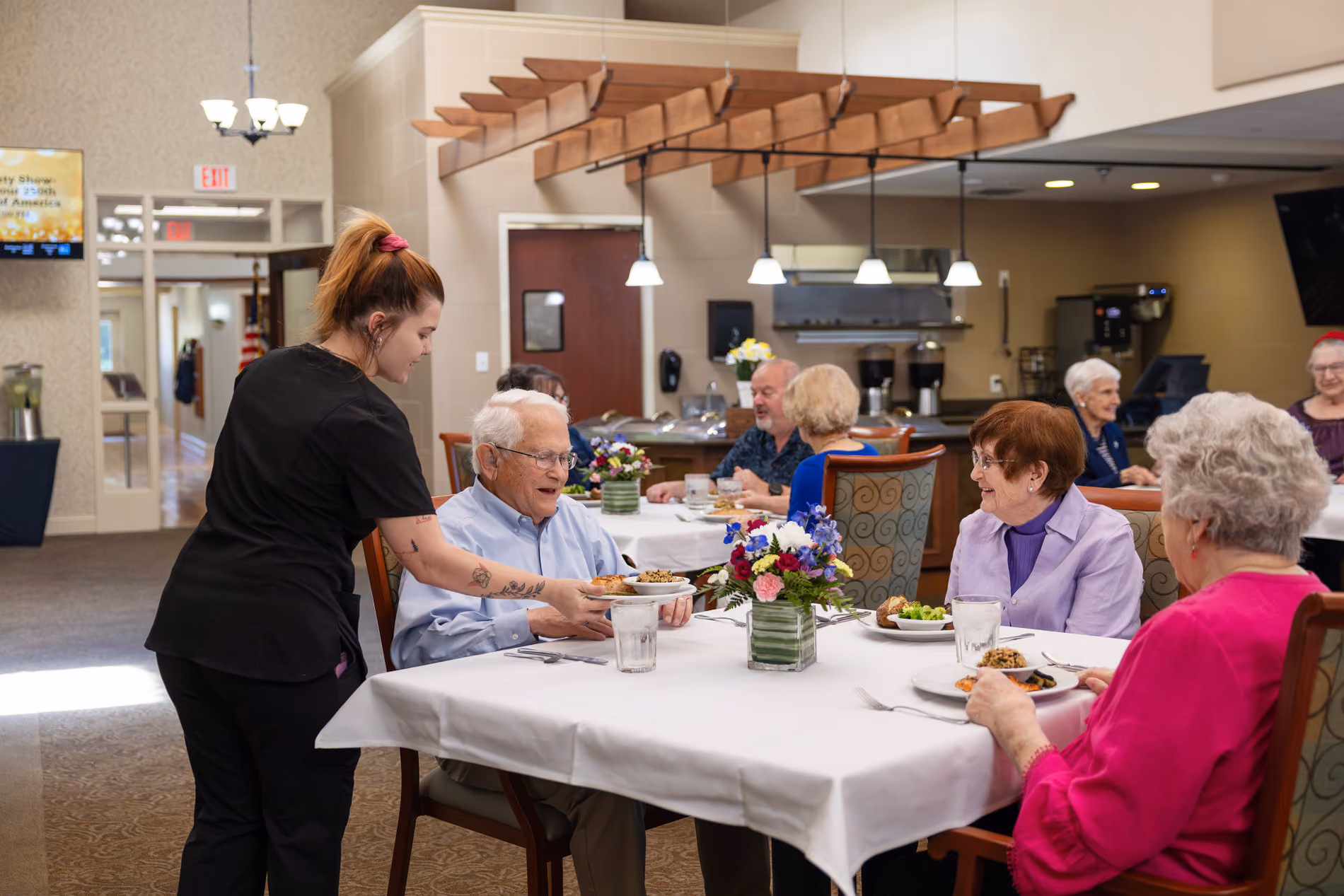 Caregiver serving meal to an elderly man while others dine and chat at a communal table.