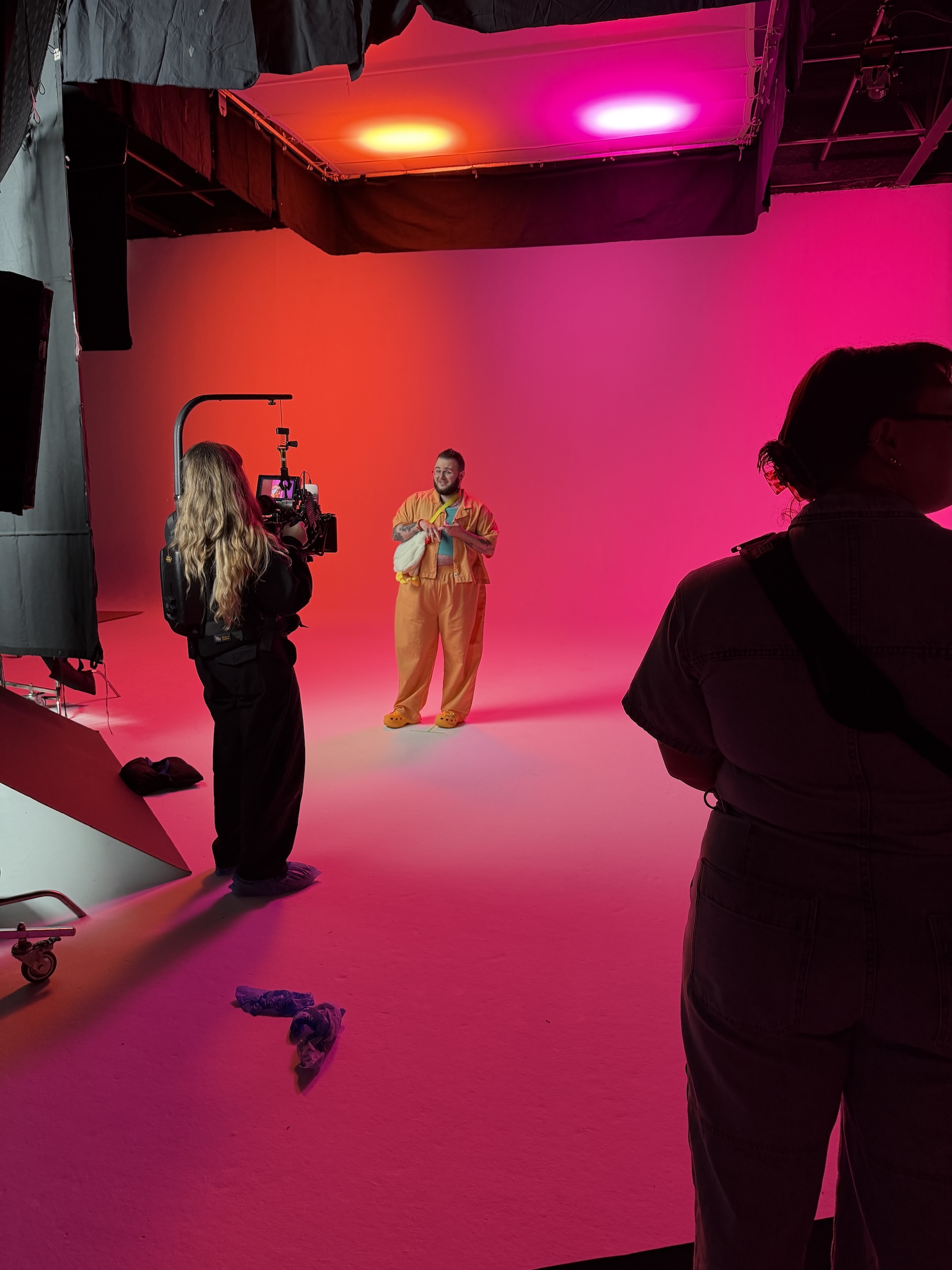 Behind the scenes at a photo shoot, with a model on a pink-lit studio set and crew silhouetted in the foreground