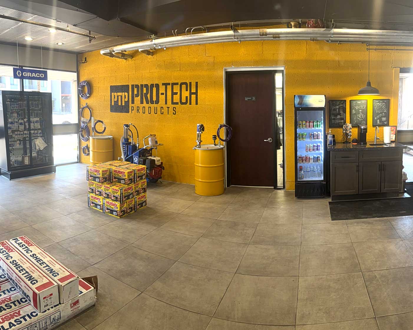 Industrial showroom with yellow barrels, branded wall, equipment, product boxes, and a refreshment area with a fridge.