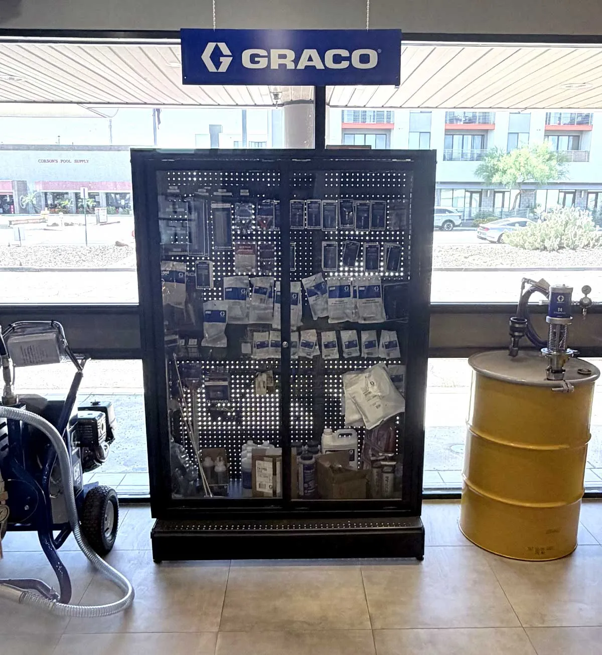 Display case with Graco branded products including hardware and fluids, between a machine and a yellow barrel.