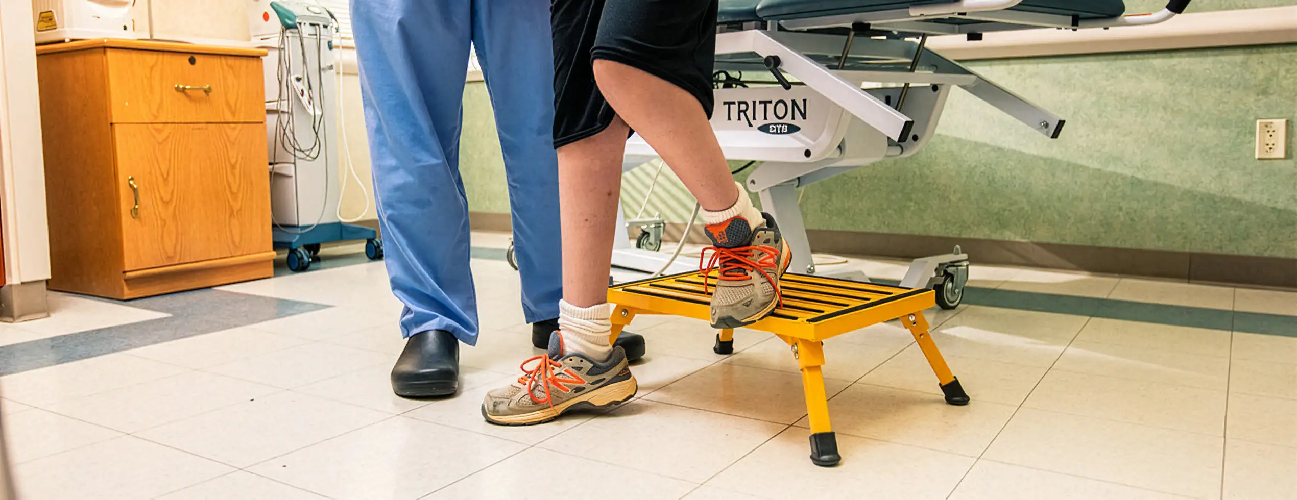 Person stepping on a yellow step stool in a medical or therapy room with a helper nearby.