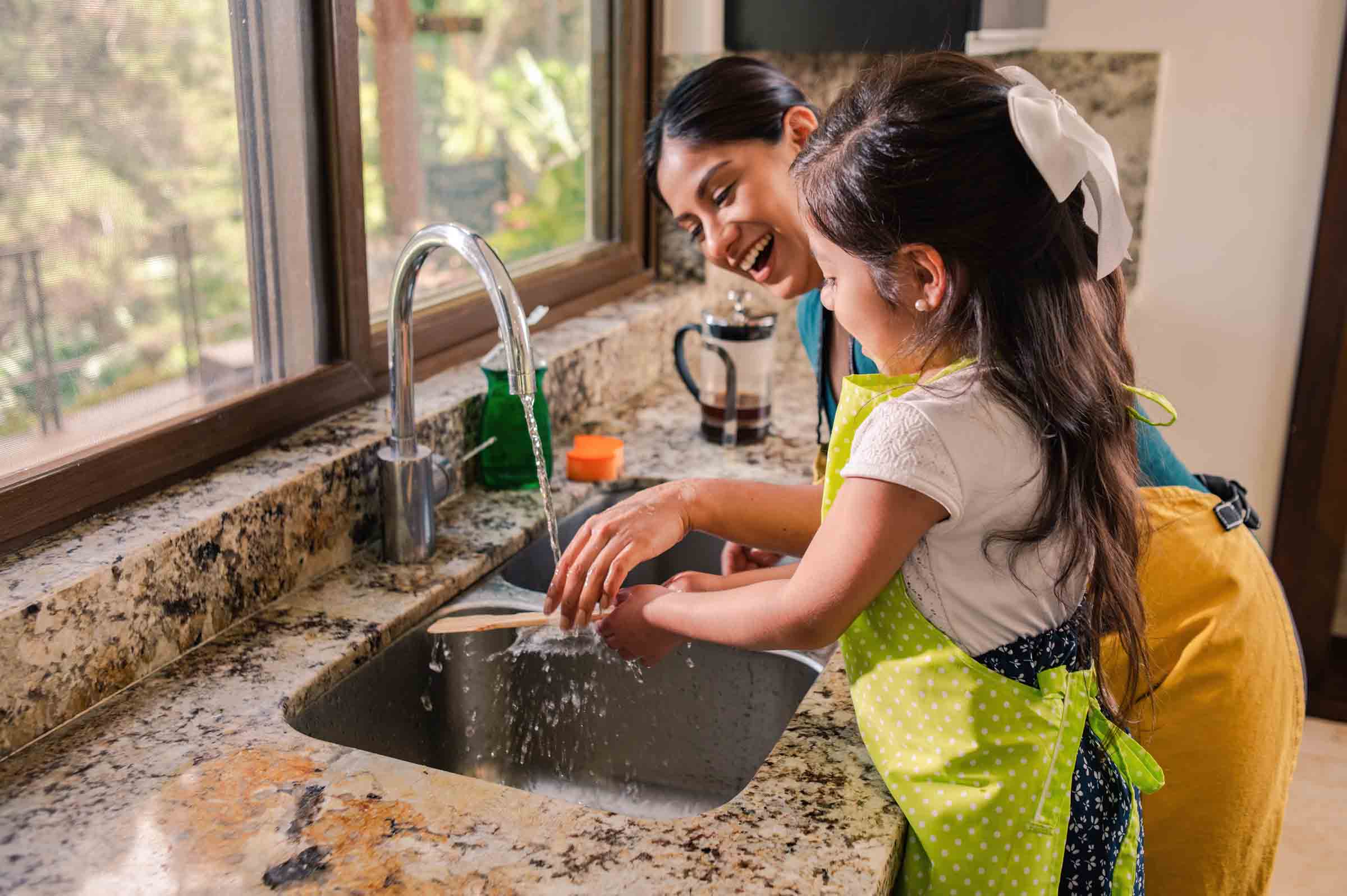 Woman and girl washing dishes together at a kitchen sink with a running faucet.