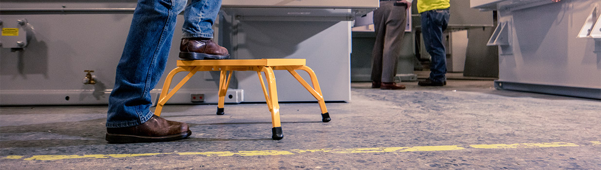 Man stepping on a small yellow stool on a factory floor with workers in the background.