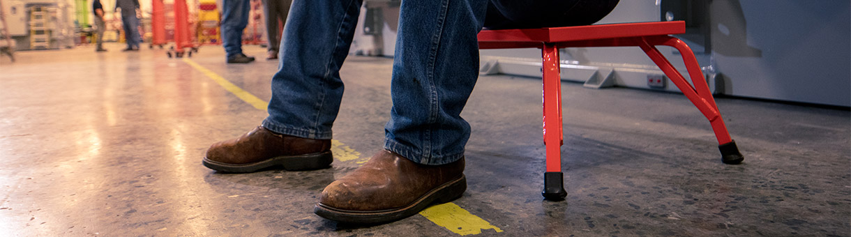 Legs in jeans and brown work boots standing on a factory floor near a red metal stool.