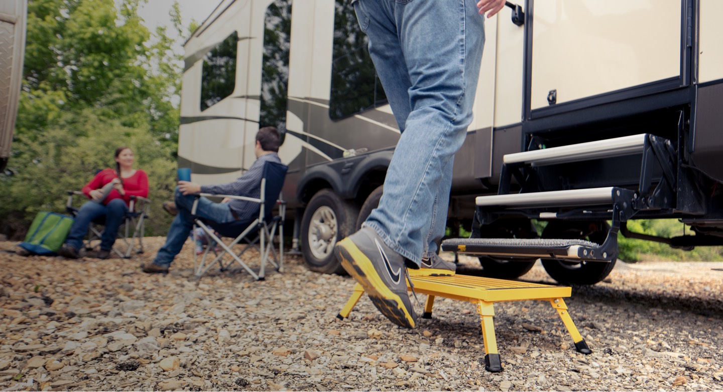 Person stepping on a yellow step stool to enter an RV with two people relaxing in camping chairs nearby.