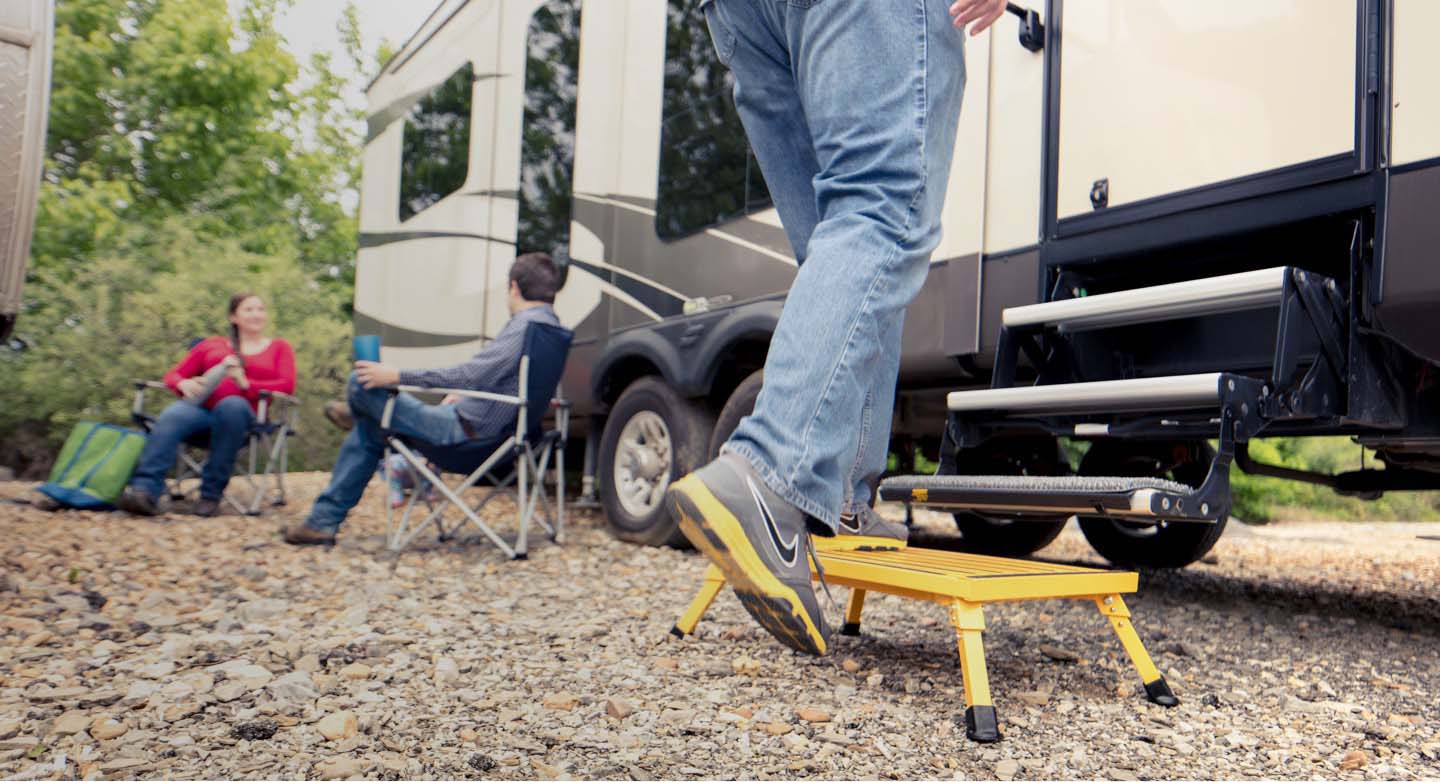 Person stepping on a yellow step stool to enter an RV while two others sit on camping chairs nearby.