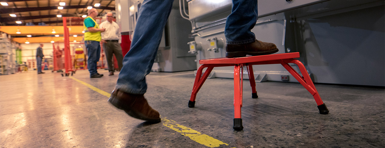 Person stepping onto a red step stool inside an industrial workshop with others in the background.