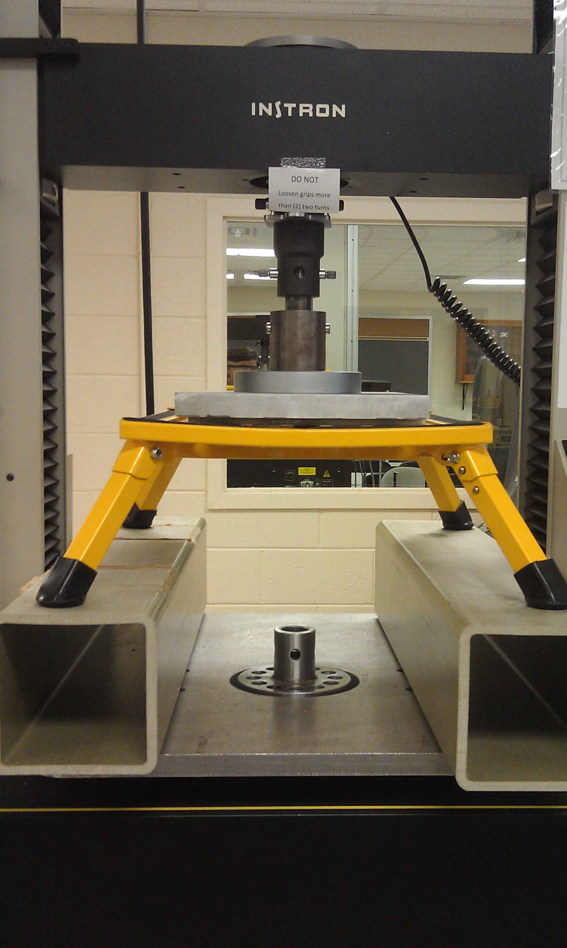 Instron testing machine with yellow stand and metal components inside a lab room.