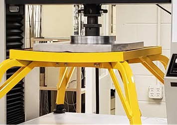 Close-up of a yellow frame supporting a metal plate under a mechanical press in a lab setting.
