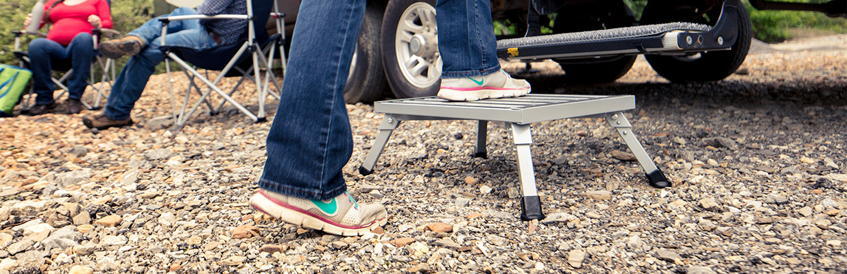 Person stepping on a small metal step stool on rocky ground near a vehicle and seated people.