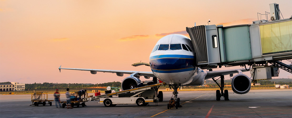 Airplane at airport gate with jet bridge and ground crew loading luggage at sunset.