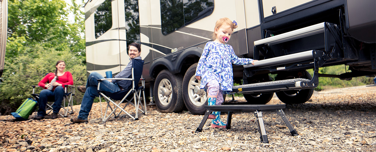 Toddler with pacifier stepping on RV entry stool while two adults sit in camping chairs nearby.