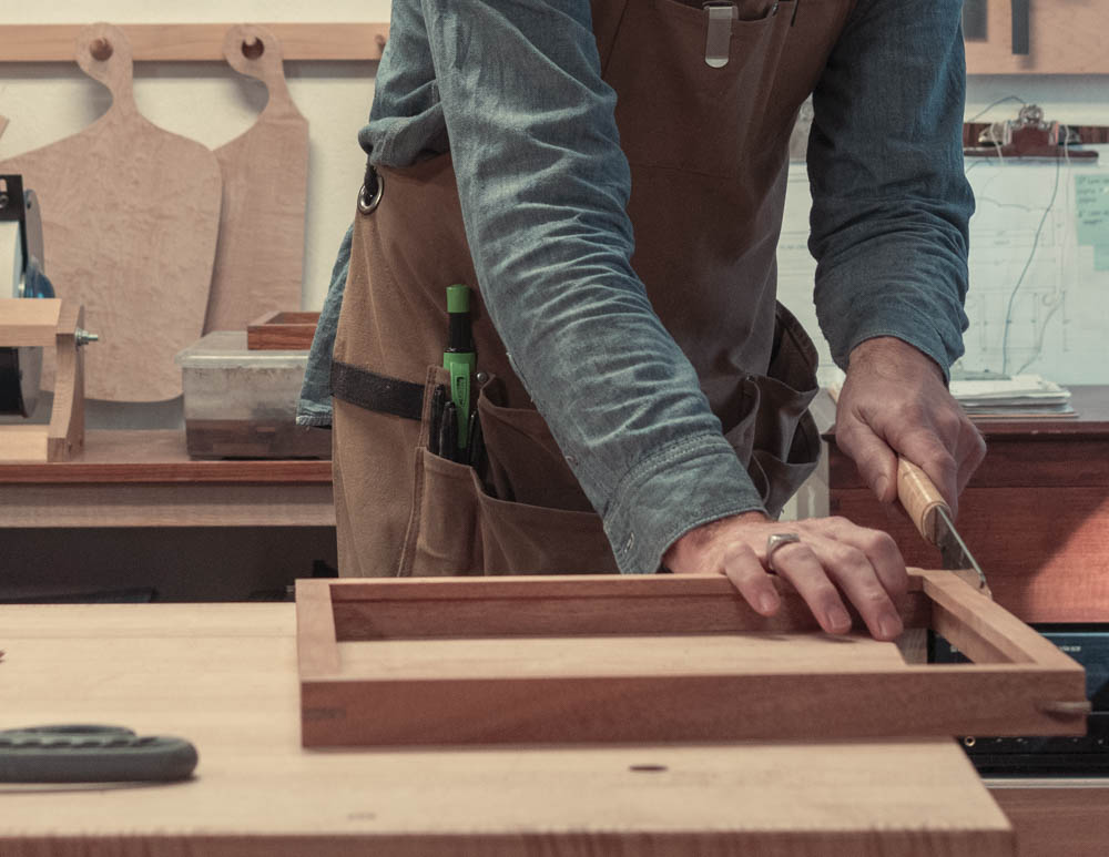 Craftsman in an apron cutting wood frame with a hand saw on a workbench.