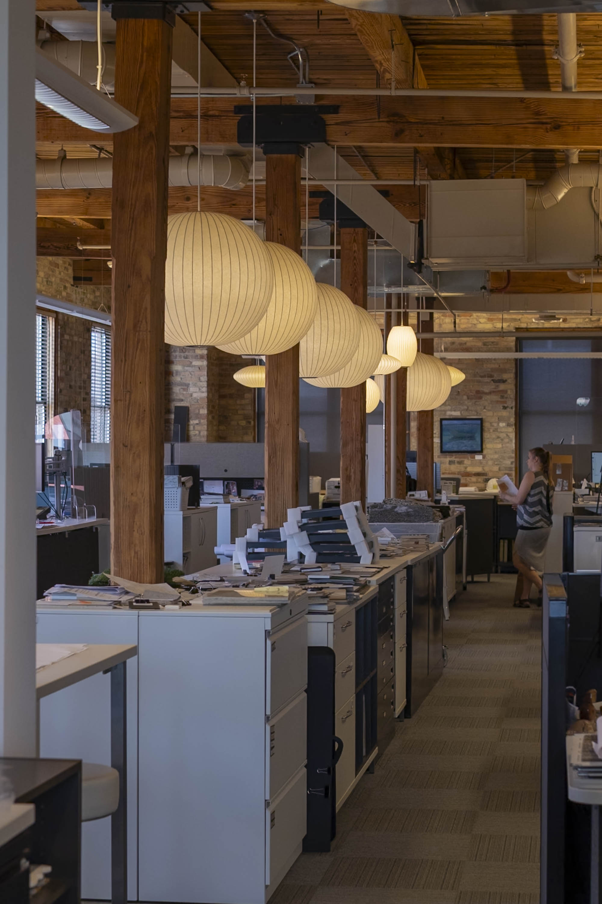 Interior office with wooden beams, hanging round paper lamps, desks, and a woman standing with papers.