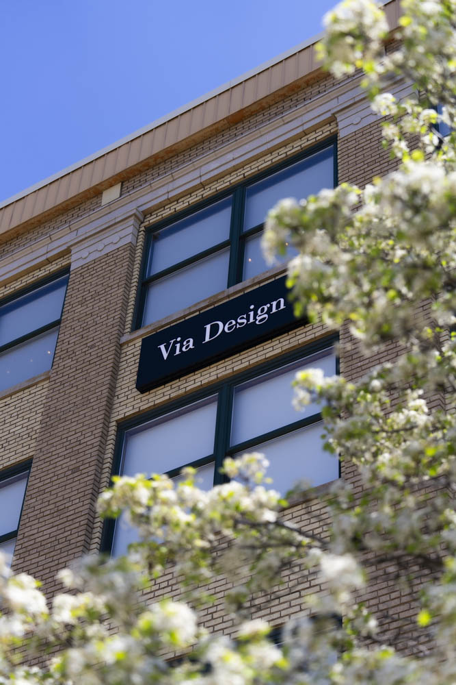 Brick office building with large windows and a sign reading 'Via Design' amid blossoming tree branches.