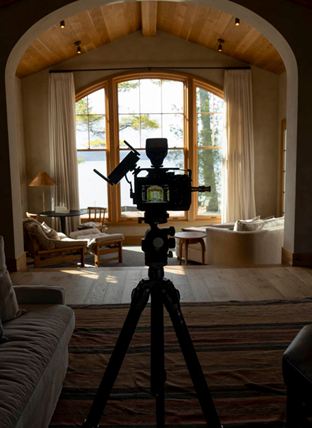 Camera on tripod in dim room facing large window with view of lake and trees outside.