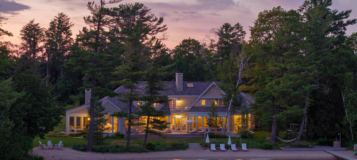 Large lakeside house surrounded by trees at sunset with lit windows, beach chairs, and a fire pit.
