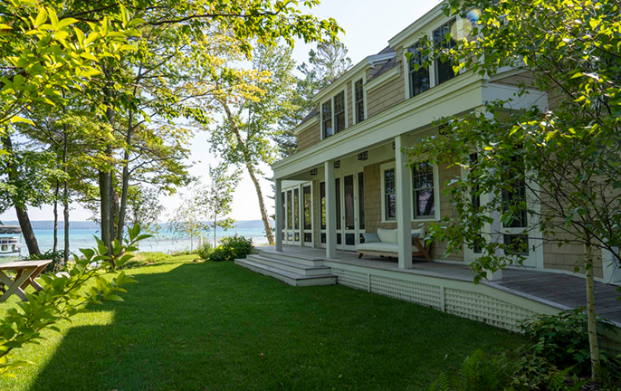 Lakeside house with large porch surrounded by green lawn and trees under blue sky.