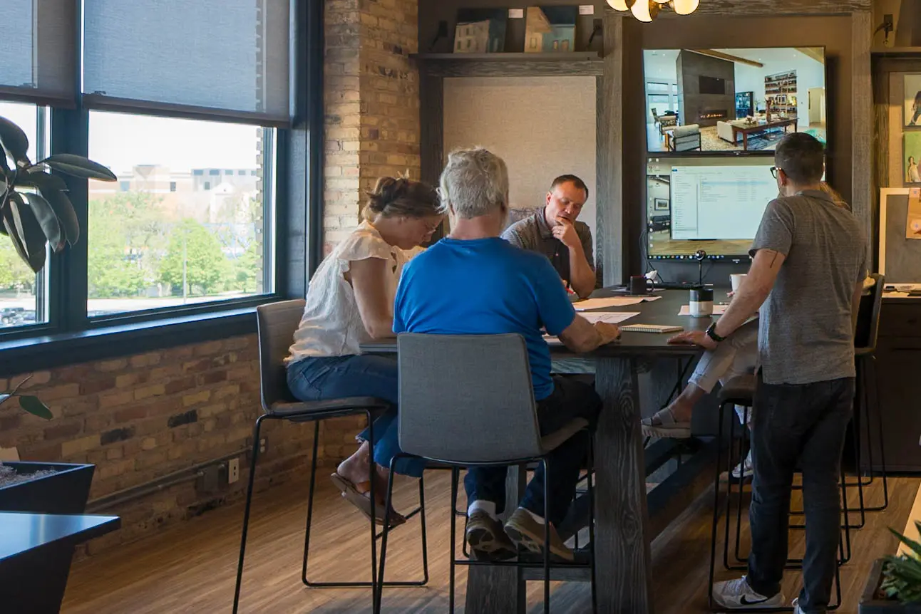 Four people in a meeting room seated and standing around a high table with two large screens on the wall.