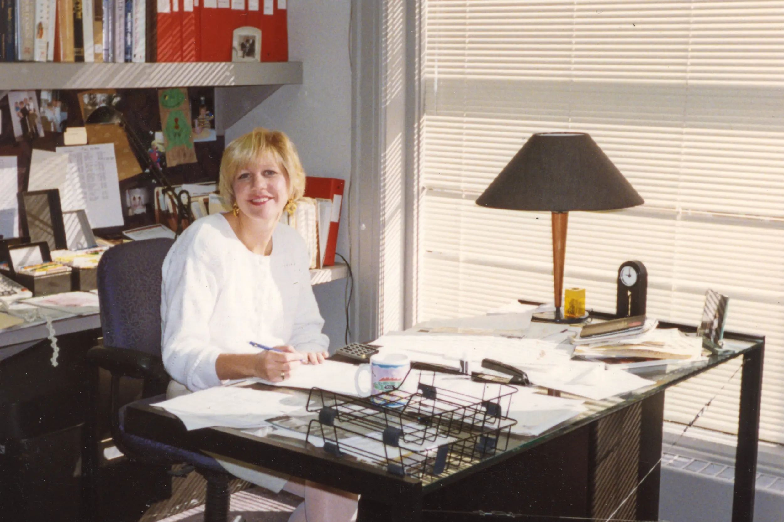 Woman in white sweater sitting at a cluttered desk with papers, lamp, and office supplies near window blinds.