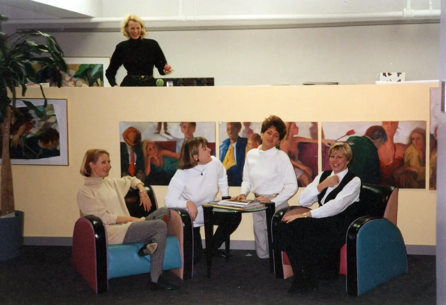 Four women seated around a small table in a room with colorful paintings and a plant, a fifth woman is standing.