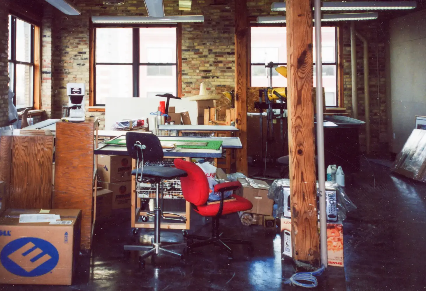 Cluttered industrial office space with red chair, desks, wooden pillars, boxes, and brick walls.