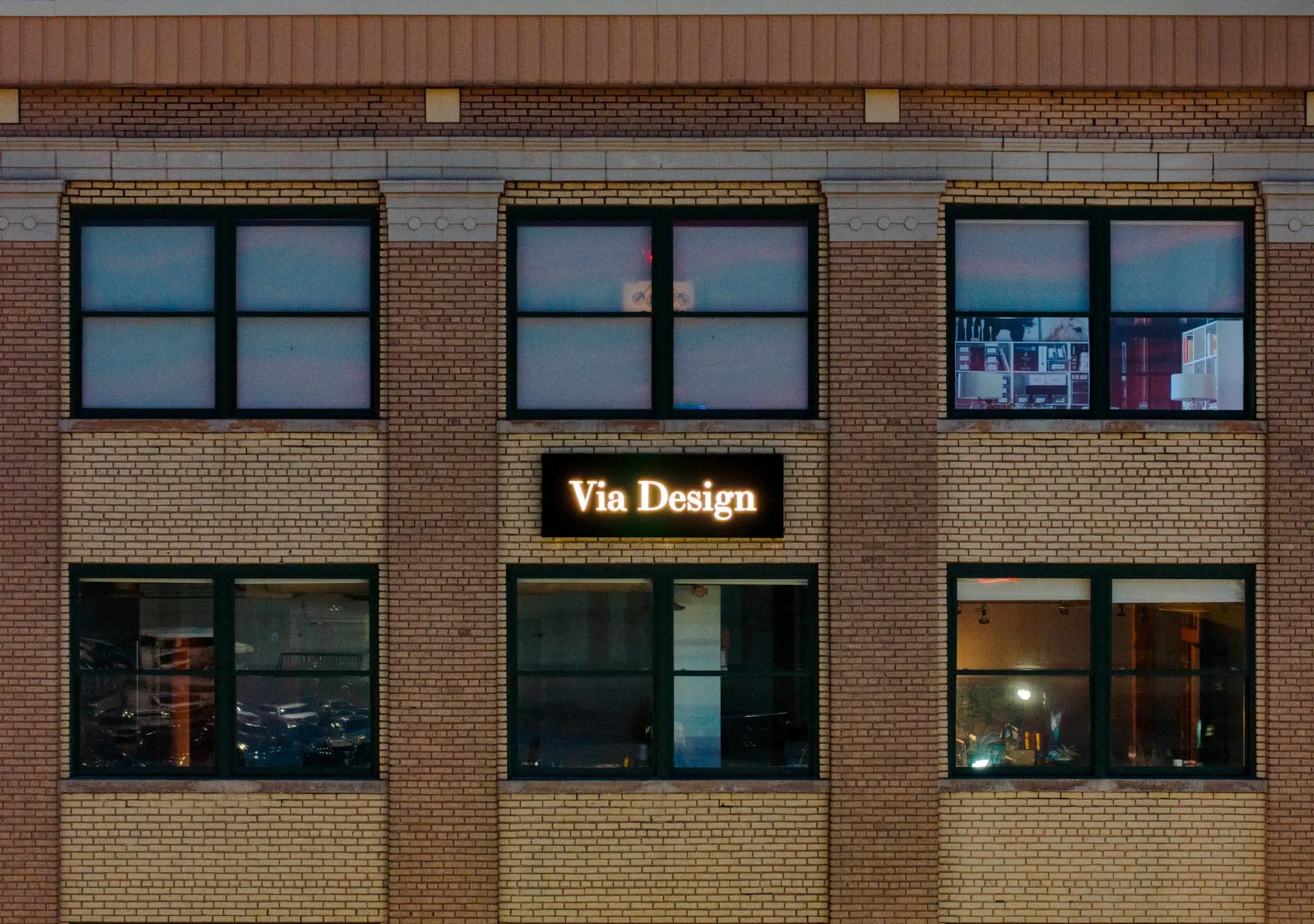 Brick building facade with black-framed windows and a lit sign reading 'Via Design' above a door.
