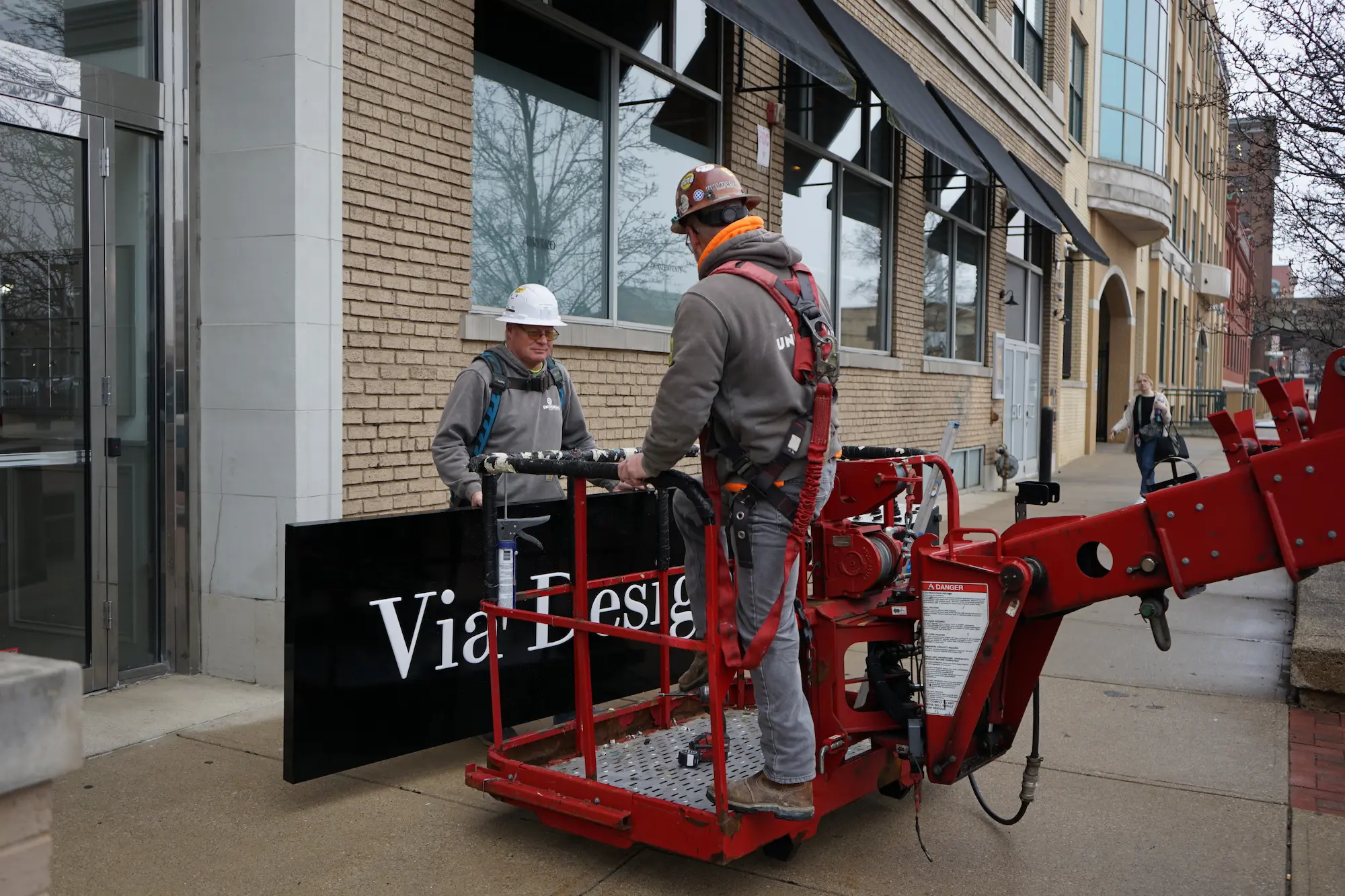 Two workers in safety gear installing a black sign with white text 'Via Desig' on a building exterior.