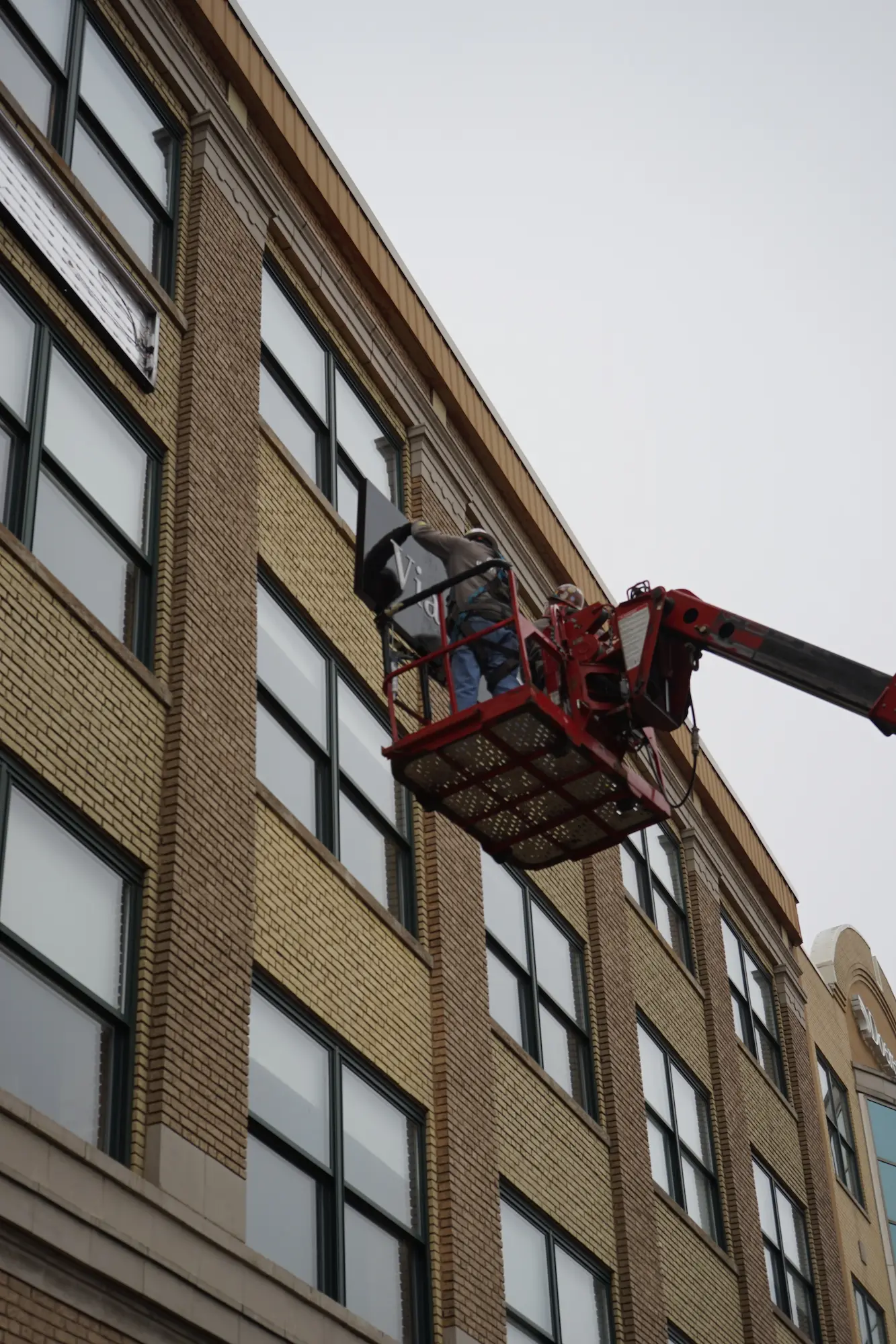 Worker on a red lift installing a sign on the side of a multi-story brick building.