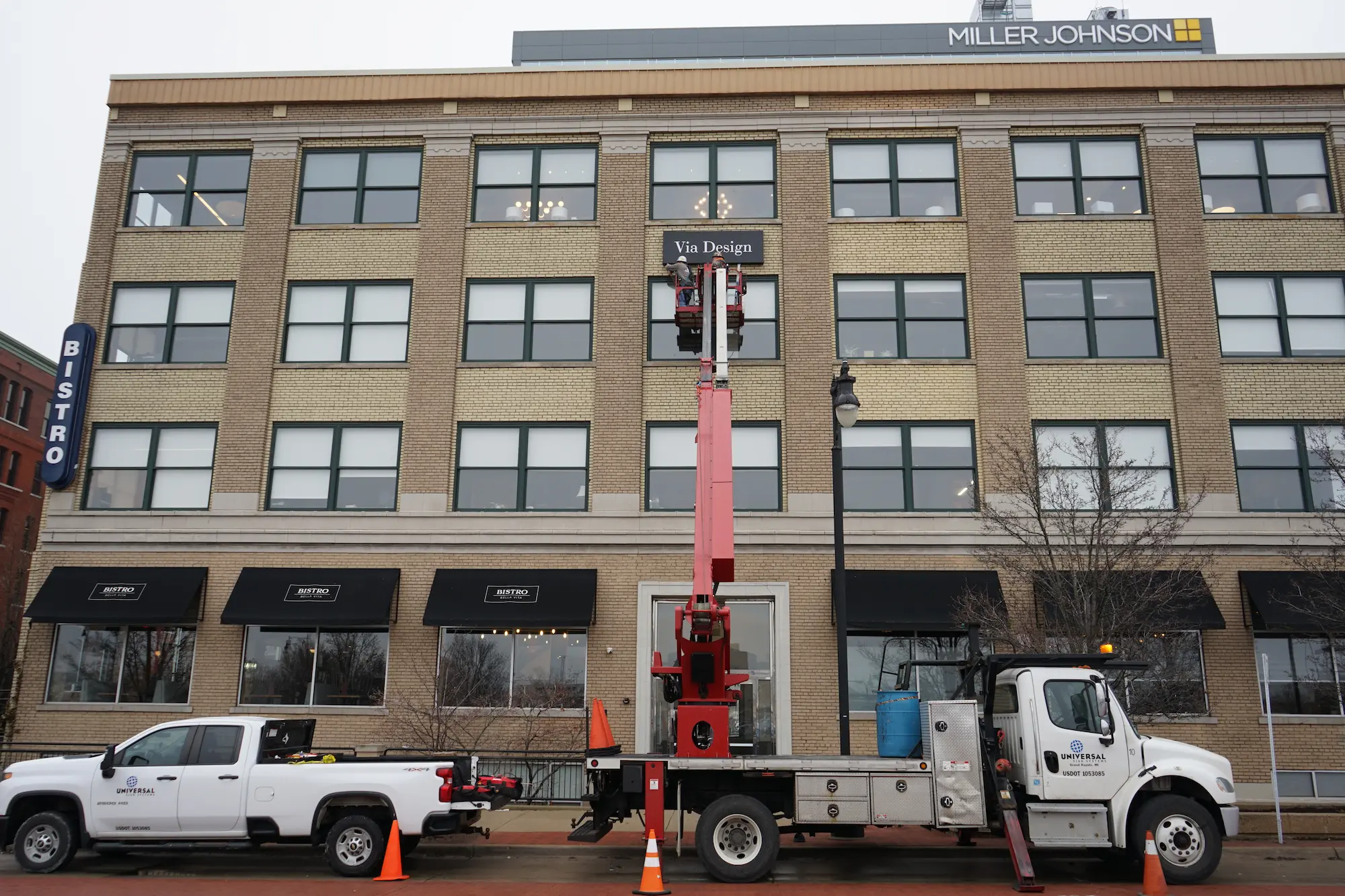 Workers on a lift installing a Via Design sign on a four-story brick building with Bistro and Miller Johnson signs.
