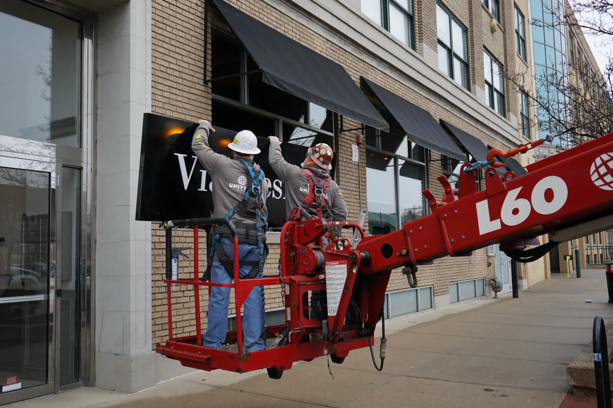 Two construction workers in safety harnesses installing a black sign on a brick building using a red lift.