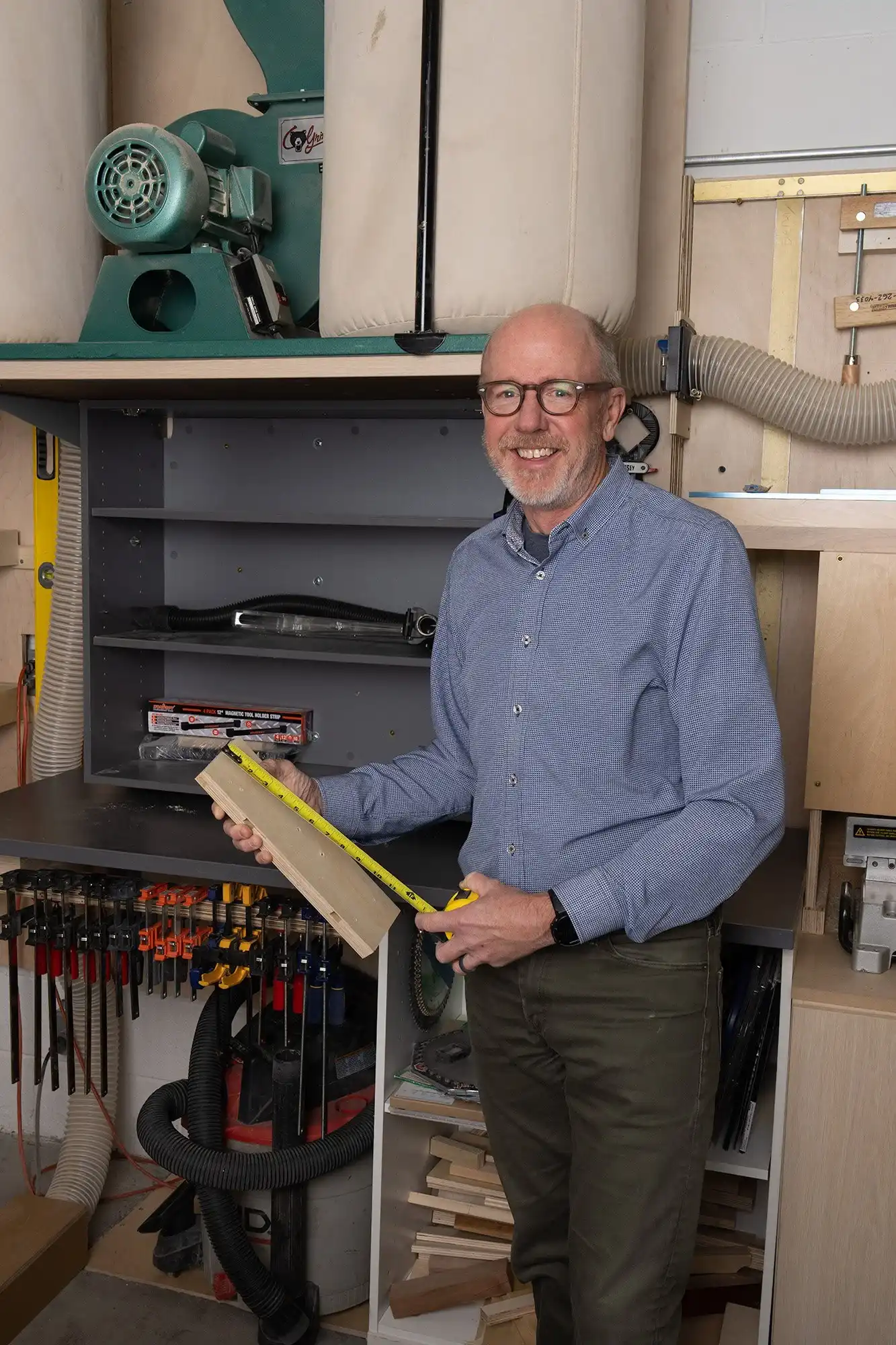 Man wearing glasses and a blue shirt measuring a wooden plank with a tape measure in a workshop.
