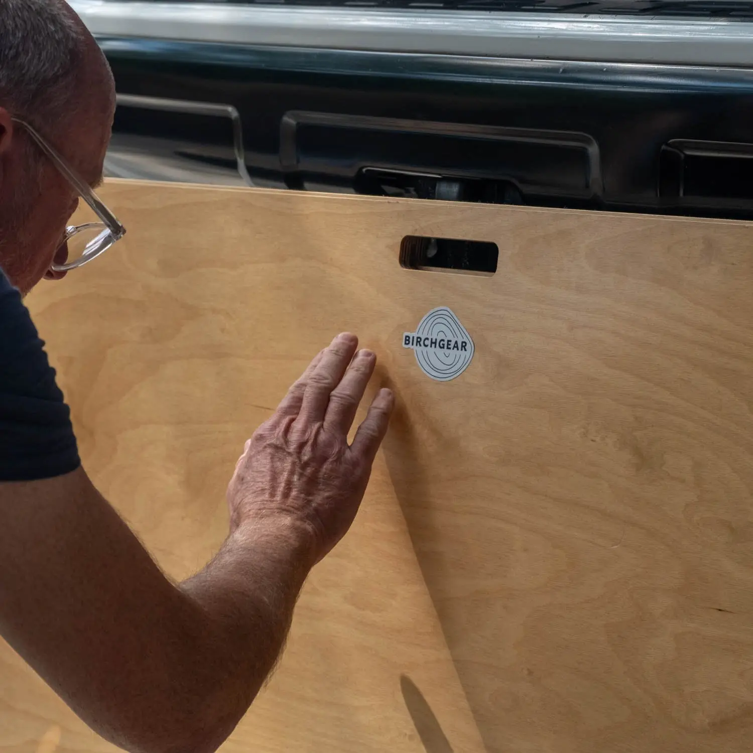 Man with glasses reaching out to touch a wooden panel with a BirchGear logo and handle cutout.