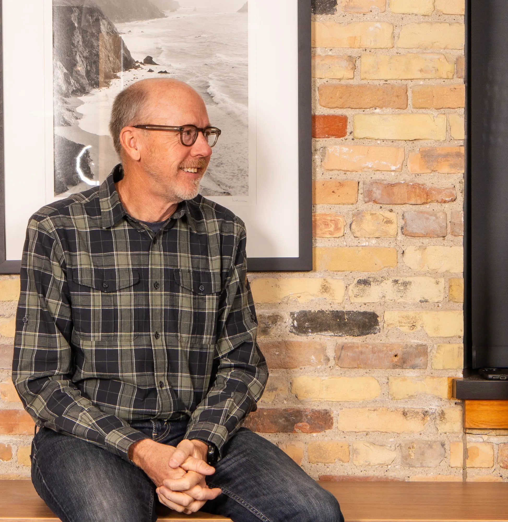 Smiling man with glasses sitting in front of a brick wall and framed photo of a coastal landscape.