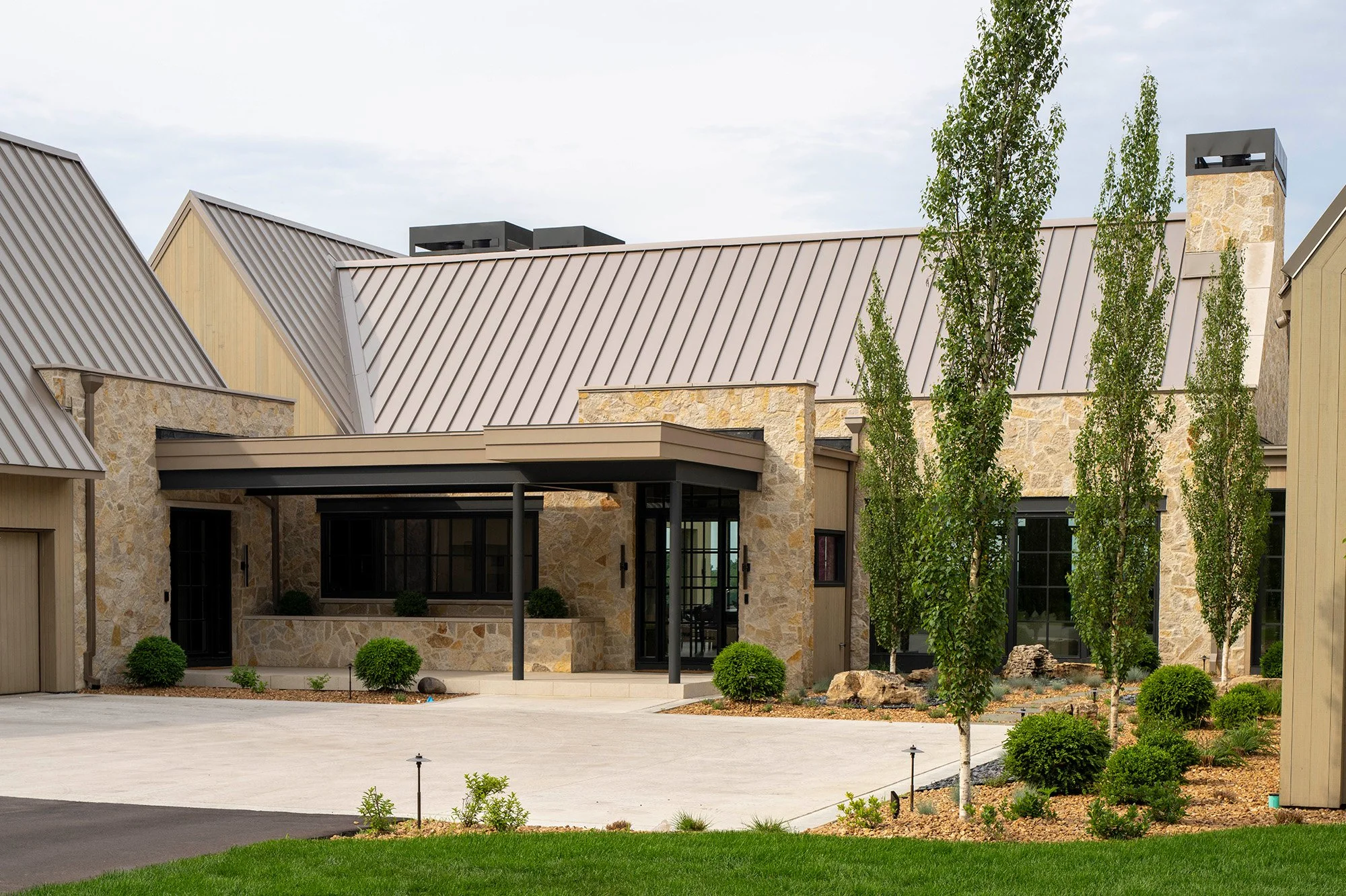 Modern house with stone walls, metal roof, tall narrow trees, and landscaped front yard.