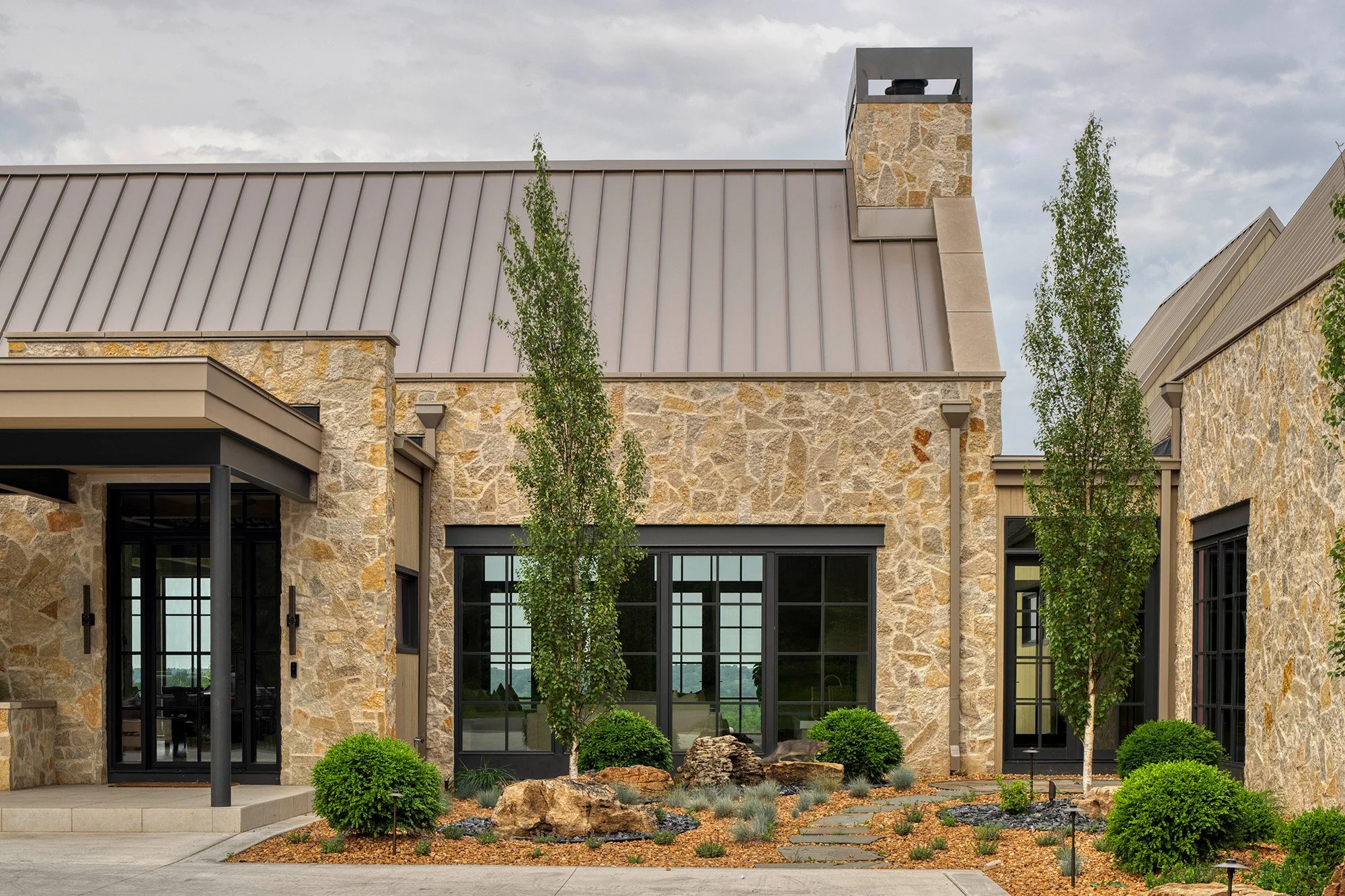 Stone house with tall windows, chimney, metal roof, and landscaped yard with trees and shrubs.