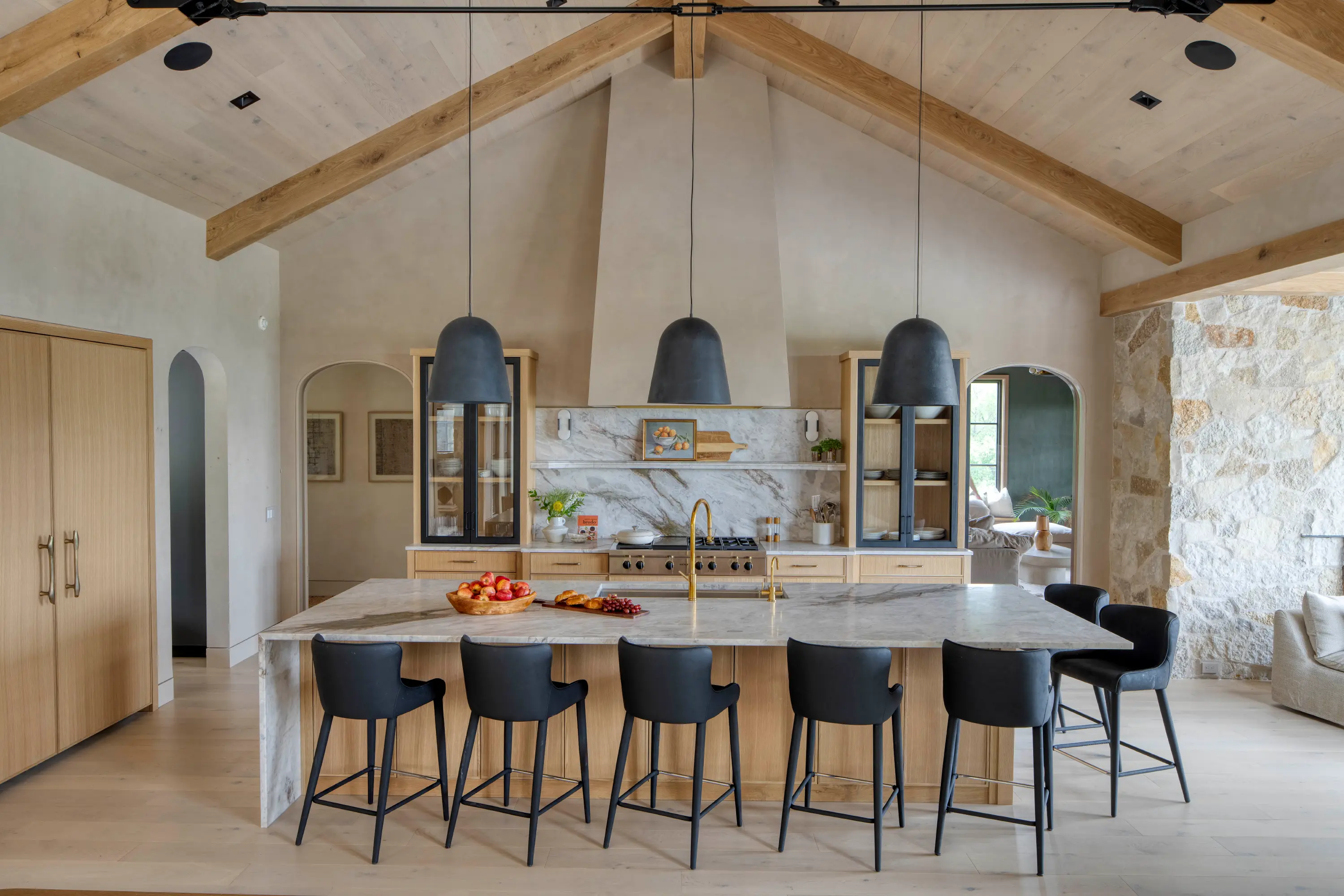 Modern kitchen with a large marble island, six black chairs, wooden cabinets, and three pendant lights.