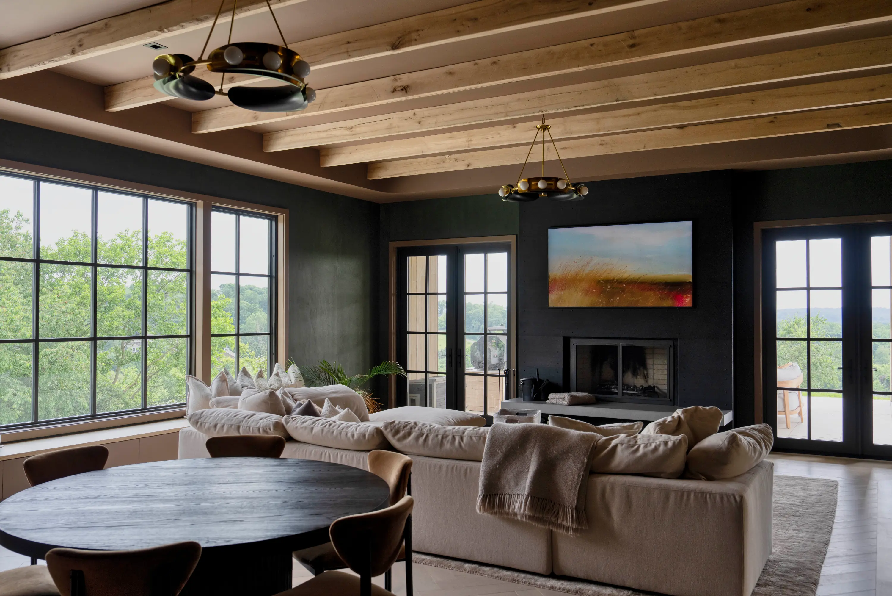 Modern living room with large windows, beige sectional sofa, fireplace, and wooden ceiling beams.