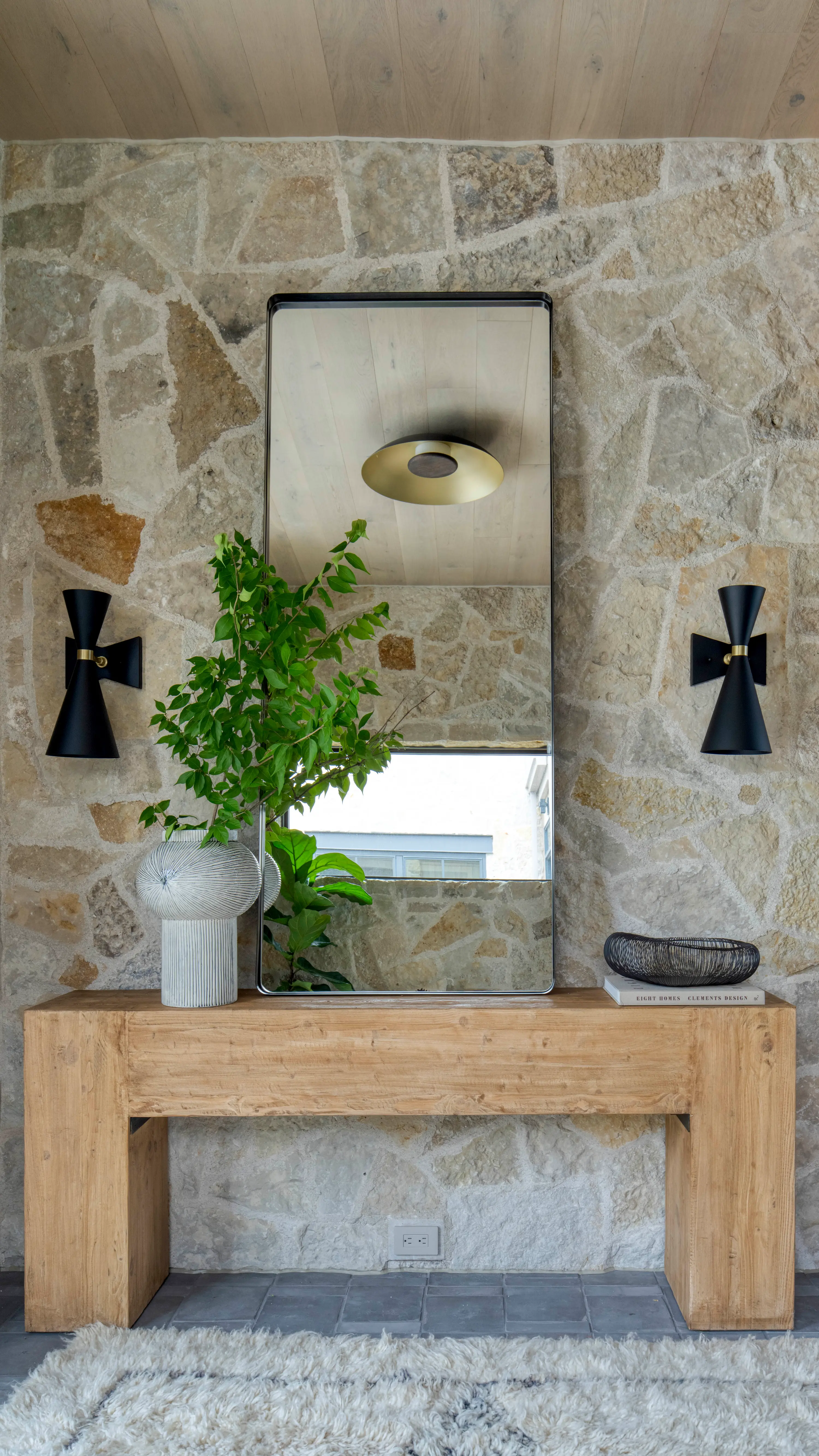 Wooden console table with a large mirror, a vase with green leaves, black wall sconces, and a decorative bowl.