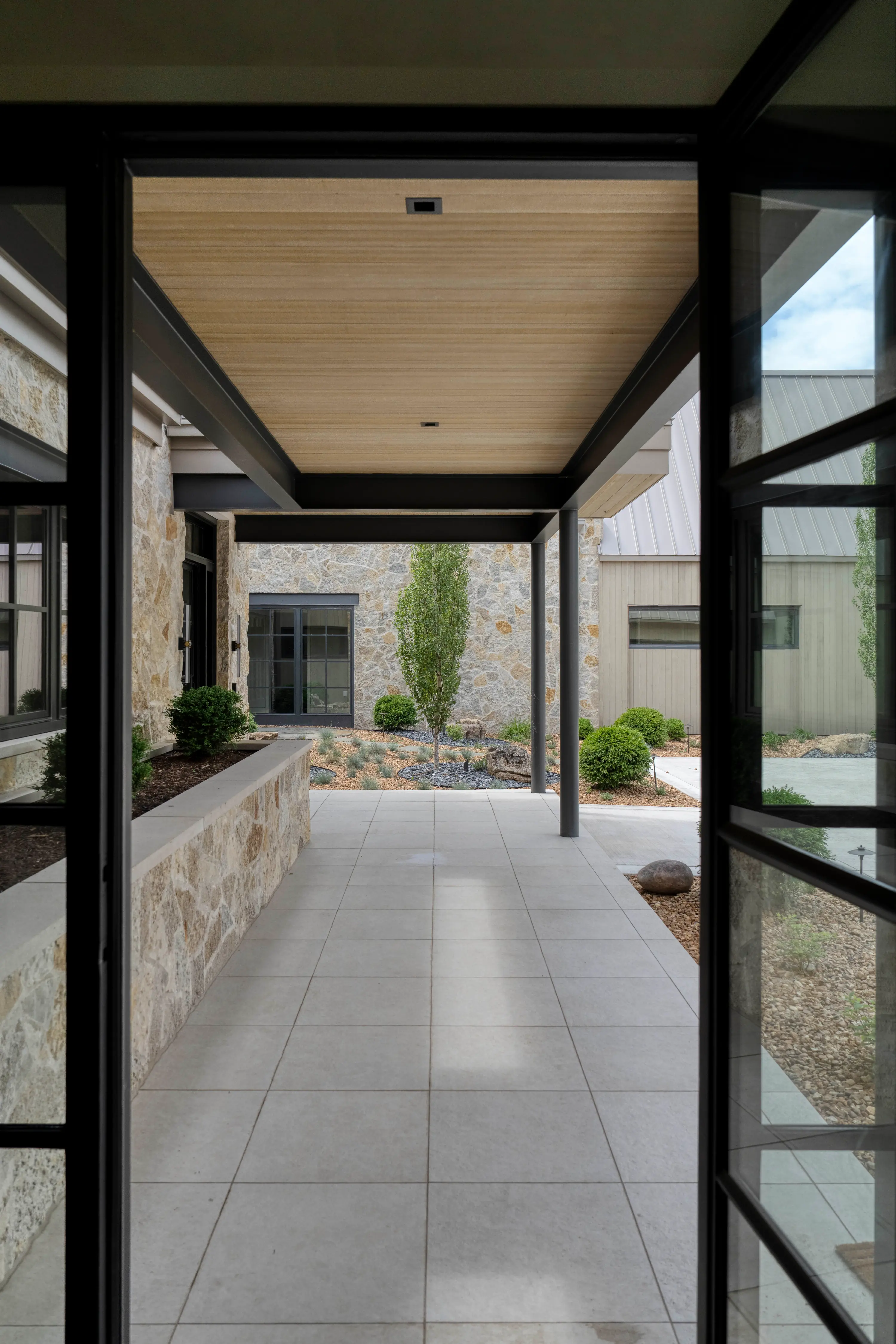 Covered stone patio with tiled floor, modern columns, shrubbery, and stone building facade.