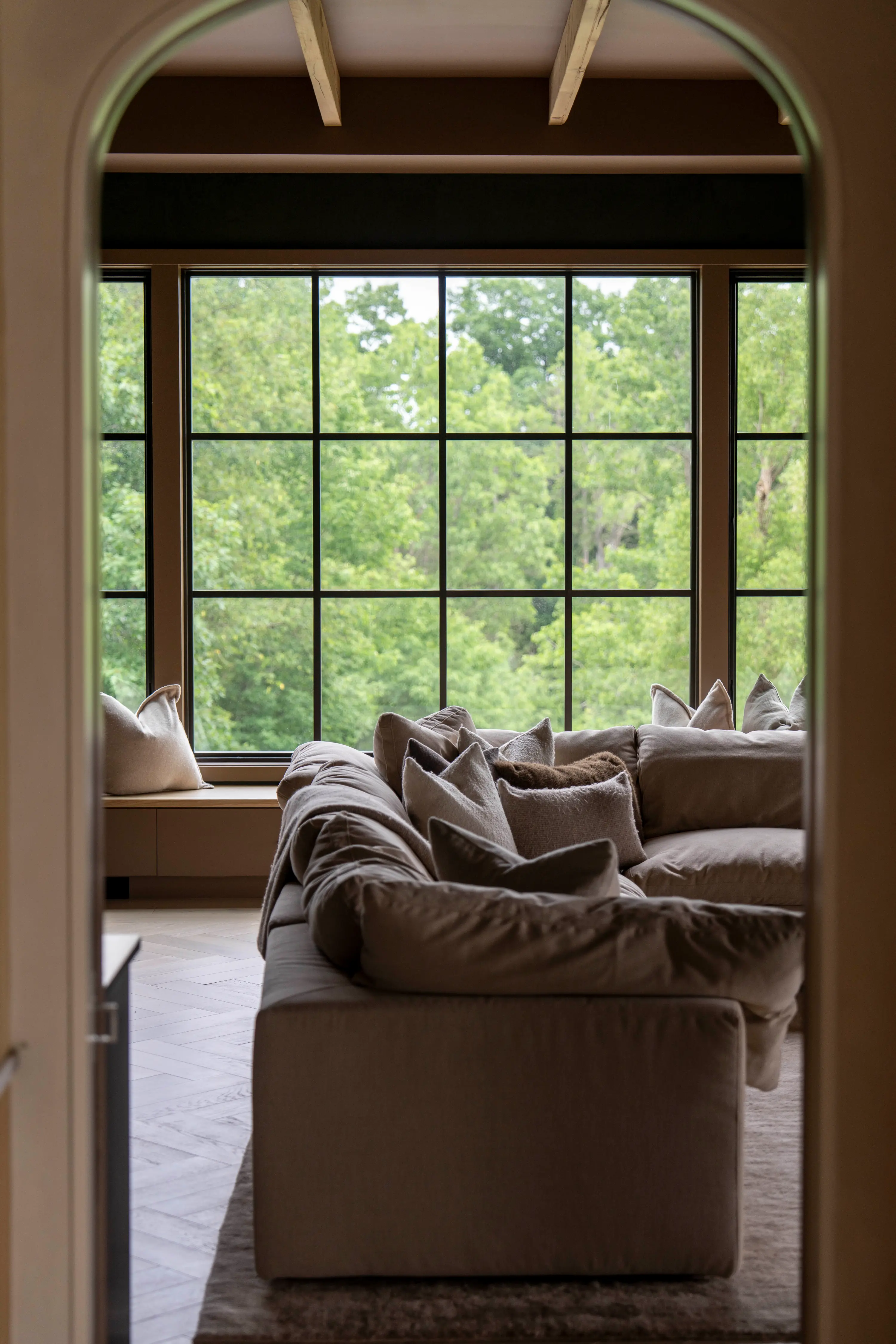 Neutral-toned sofa with pillows in front of large window showing green leafy trees outside.