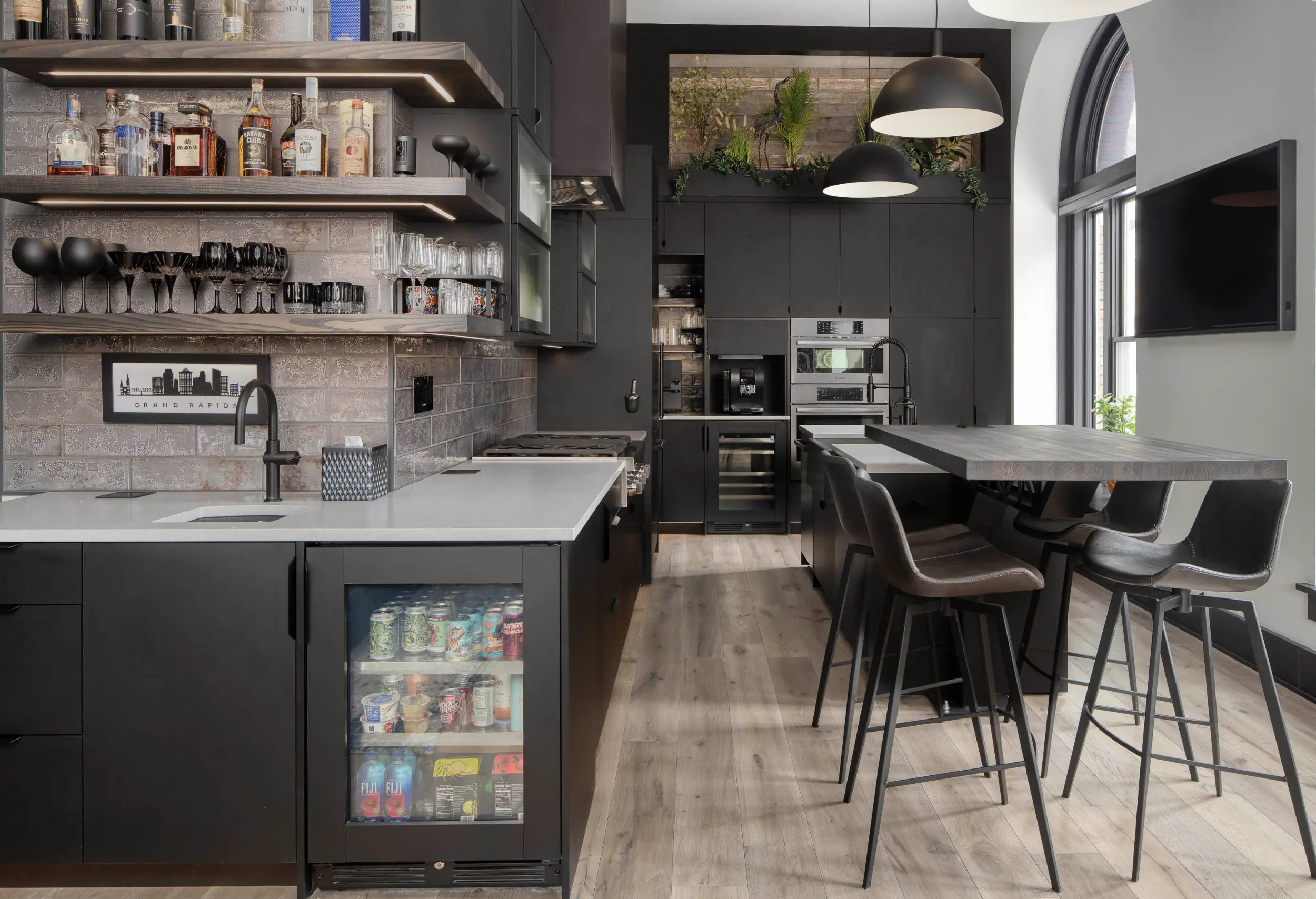 Modern kitchen with black cabinets, bar stools, arched window, beverage fridge, and hanging pendant lights.