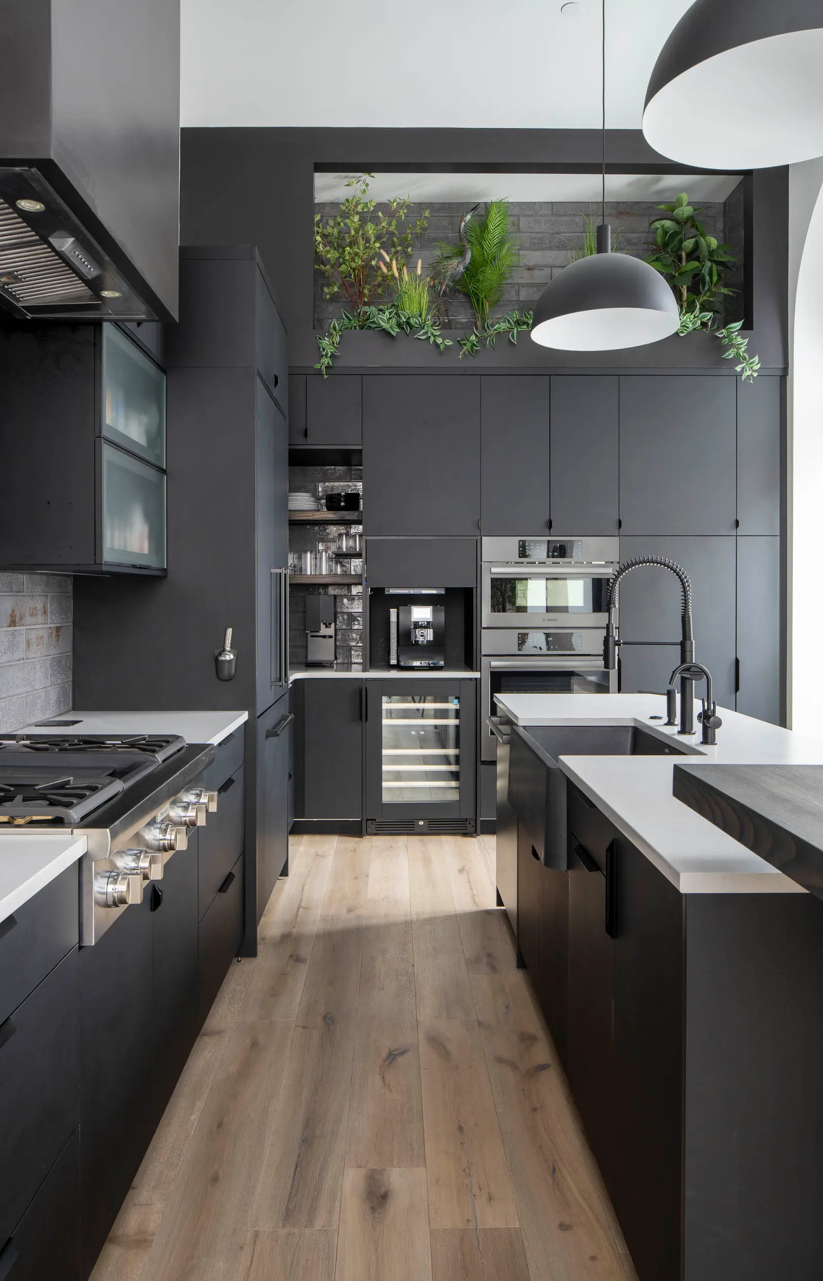 Modern kitchen with dark cabinets, white countertops, built-in ovens, and plants above cabinets.