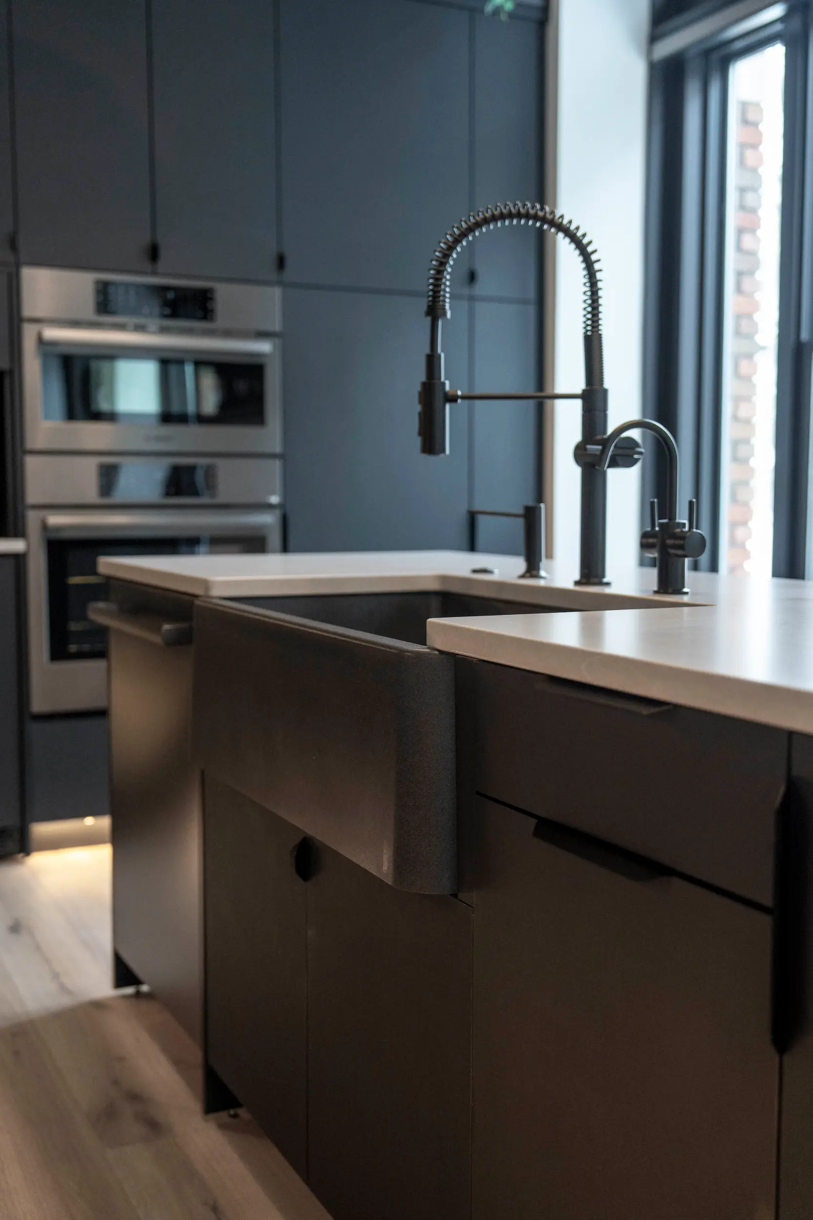 Modern kitchen with dark cabinetry, a farmhouse sink, a flexible spring coil faucet, and built-in stainless steel ovens.