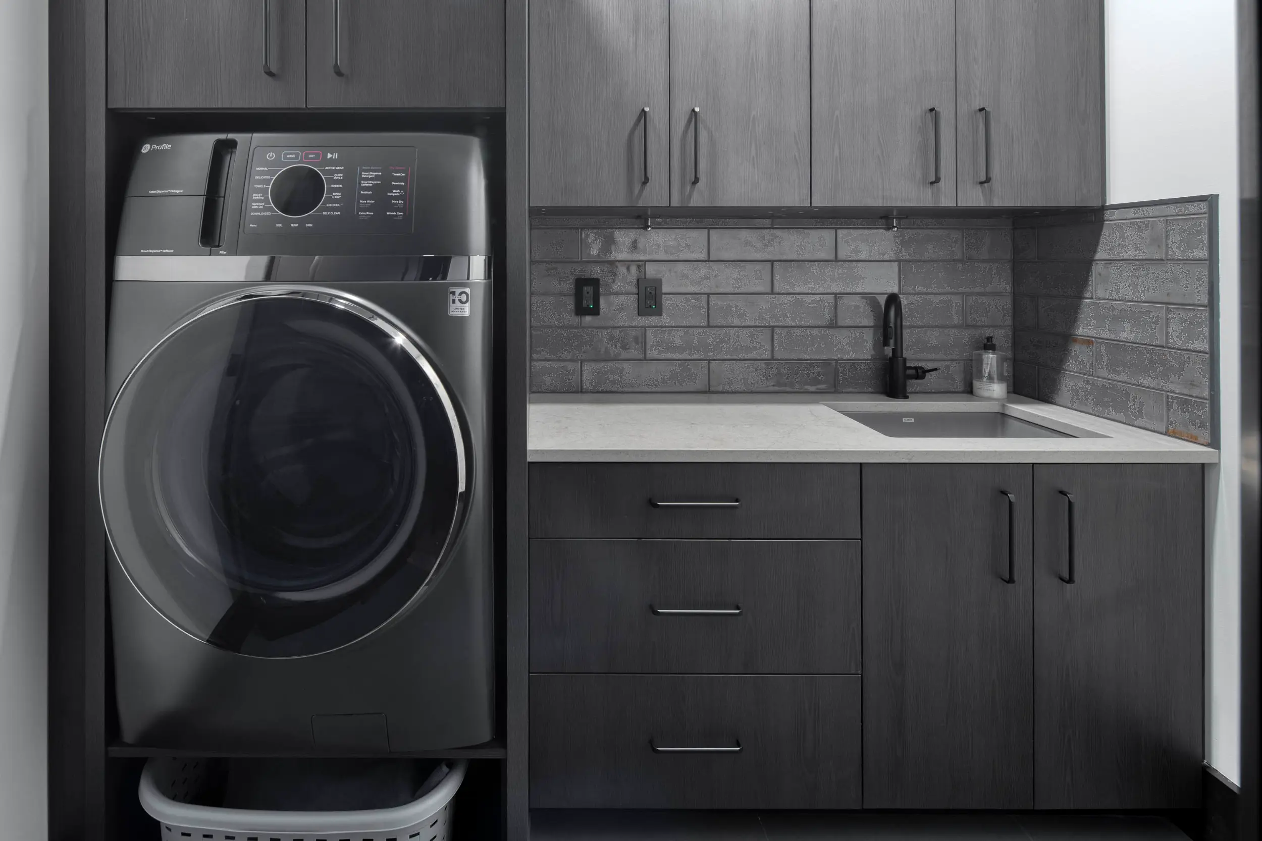 Modern laundry room with dark wood cabinets, front-loading washer, white countertop, sink, and gray tiled backsplash.