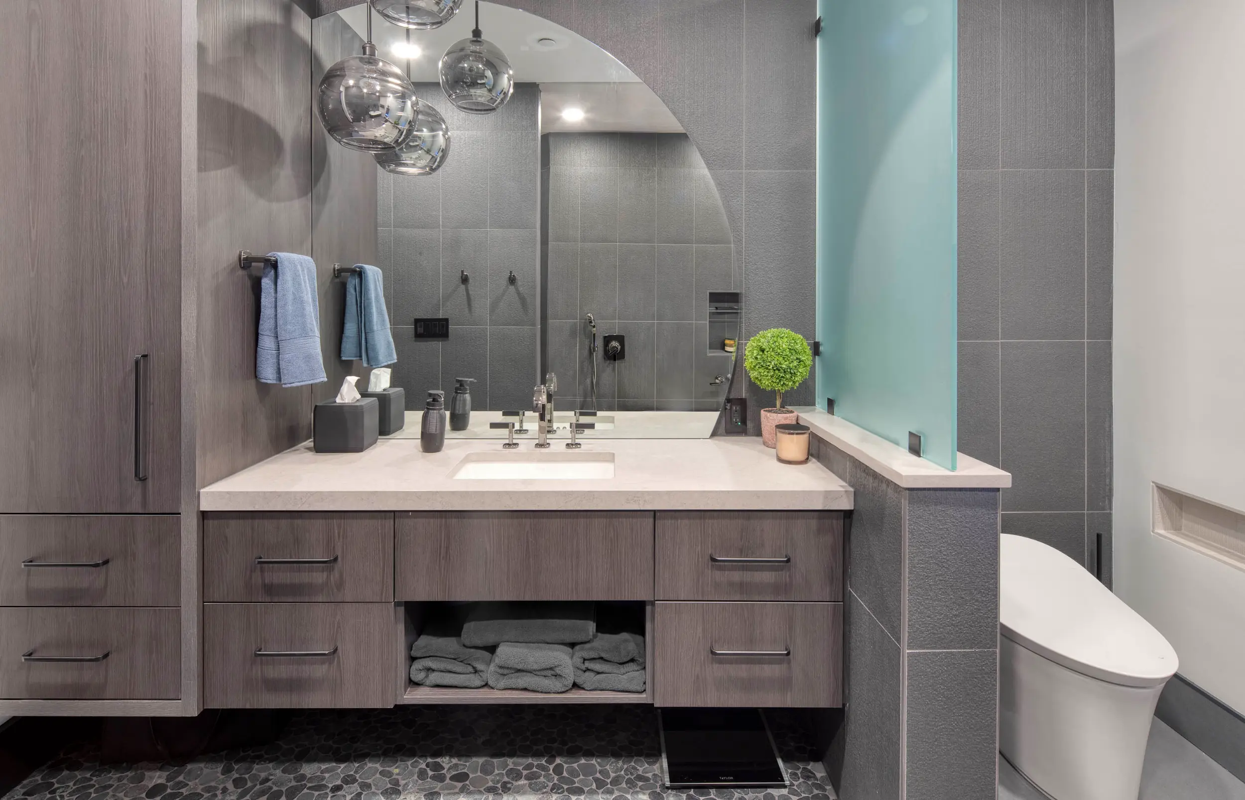Modern bathroom vanity with gray wood cabinets, large round mirror, stone countertop, and frosted glass divider.