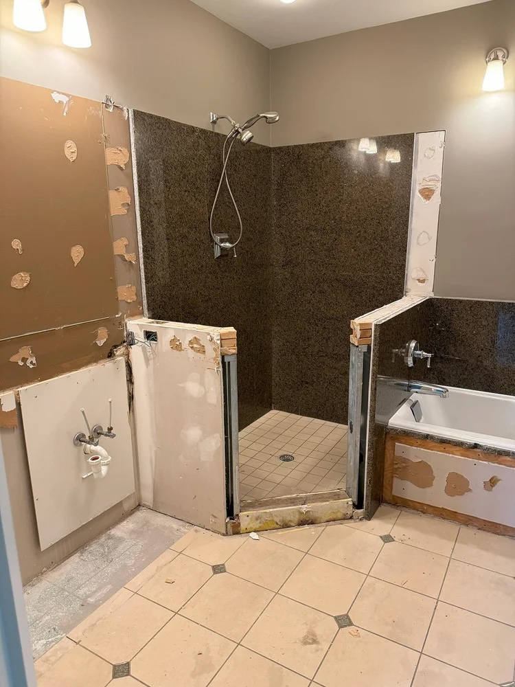 Partially renovated bathroom with tiled floor, corner shower with dark walls, and bathtub with exposed panel.